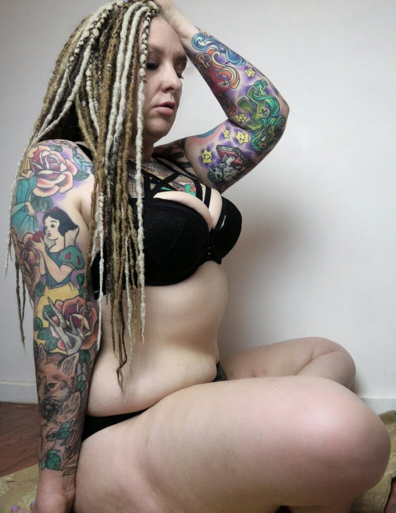 Rem Sequence alternative tattooed adult model from Australia with sleeve and chest tattoo and dreadlocks wearing black lingerie and sitting cross legged on the floor in profile