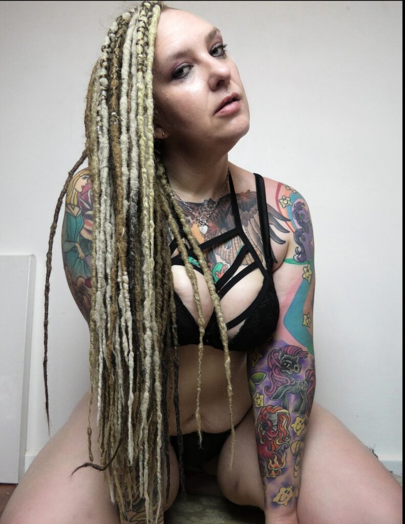 Rem Sequence tattooed Aussie adult model with dreadlocks wearing black lingerie and leaning forward toward camera