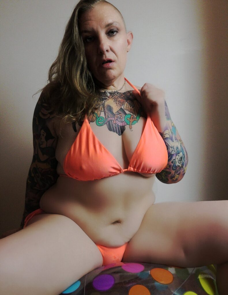 Rem Sequence alternative tattooed adult model from Australia sitting on a polka dot beach ball and wearing a fluoro orange bikini with blond hair loose on her shoulders